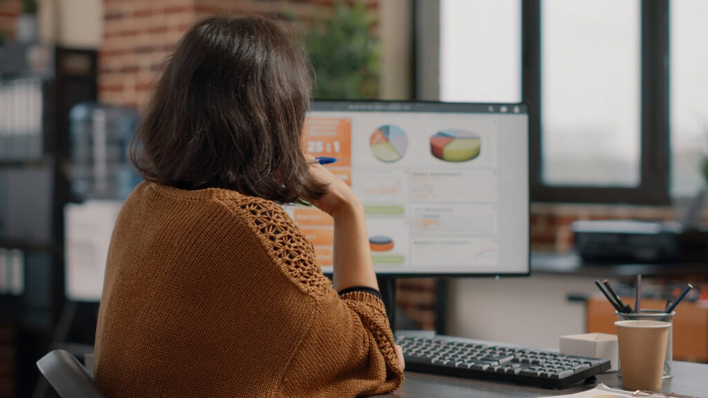 Das Foto zeigt eine Geschäftsfrau in einer Marketingagentur, die mit der Analyse von Daten zur Optimierung der Customer Journey beschäftigt ist. Sie ist von hinten aufgenommen und blickt auf einen Computerbildschirm, auf dem verschiedene farbige Datenvisualisierungen angezeigt werden, darunter Torten- und Liniendiagramme, die typische Instrumente zur Analyse der Unternehmensleistung und der Kundenbindung sind. Die Frau hält einen Stift in der Hand, was darauf hindeutet, dass sie aktiv mit den Daten arbeitet. Sie sitzt an einem Schreibtisch mit einer Tastatur vor sich, neben einer Kaffeetasse aus Papier steht eine Tasse mit mehreren Stiften und Bleistiften, was auf eine Arbeitsumgebung hinweist. Die Umgebung ist gemütlich mit einem Hauch von Modernität, unterstrichen durch die freiliegende Ziegelwand und die großen Fenster, durch die natürliches Licht in den Raum fällt. Die Kleidung und die Körperhaltung spiegeln eine professionelle und konzentrierte Haltung in einem modernen Büro wider.