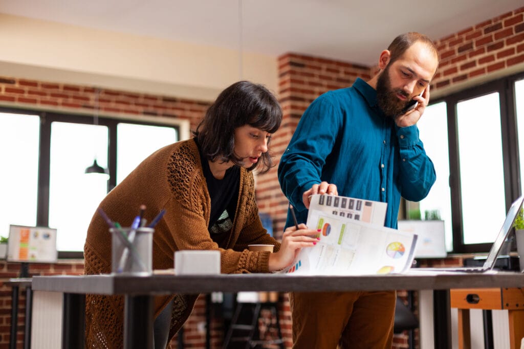 Das Foto zeigt eine Geschäftsfrau und ihren Kollegen bei der Arbeit in einer Marketingagentur. Die Frau beugt sich über ein Dokument und zeigt auf einen bestimmten Abschnitt, was darauf schließen lässt, dass sie Daten oder Strategien im Zusammenhang mit der Optimierung der Customer Journey diskutiert. Ihr Gesichtsausdruck ist konzentriert und sie scheint ihrem Kollegen den Inhalt des Dokuments zu erklären oder zu analysieren. Ihr männlicher Kollege ist gleichzeitig in ein Telefongespräch vertieft, was auf Multitasking oder die Abstimmung von Informationen mit einer dritten Person hindeutet. Auch er hält ein Dokument in der Hand, auf das er sich während des Gesprächs zu beziehen scheint. Die beiden befinden sich in einem hellen Büro mit großen Fenstern, sichtbaren Ziegelwänden und modernen Möbeln, die einen dynamischen und zeitgemäßen Arbeitsplatz widerspiegeln. Das Bild vermittelt den Eindruck aktiver Zusammenarbeit und Kommunikation, beide Personen sind in ihre Arbeit vertieft. Die legere, aber dennoch professionelle Kleidung der beiden Personen sowie die offene und gut beleuchtete Umgebung lassen auf eine angenehme und fortschrittliche Büroatmosphäre schließen.