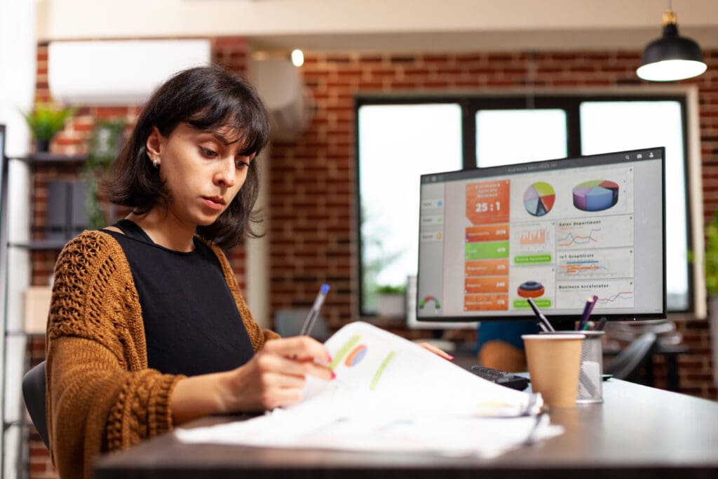 Das Foto zeigt eine Geschäftsfrau in einer Marketingagentur, die sich intensiv mit der Analyse von Daten zur Optimierung der Customer Journey beschäftigt. Sie sitzt, ihre Aufmerksamkeit ist auf ein Dokument in ihren Händen gerichtet, vielleicht studiert sie Berichte oder Erkenntnisse. Sie hält einen Stift bereit, um sich Notizen zu machen oder wichtige Daten zu markieren. Im Hintergrund zeigt ein großer Monitor ein lebendiges Dashboard mit verschiedenen Datenvisualisierungen, darunter Torten- und Balkendiagramme, die eine umfassende Analyse der Geschäftskennzahlen suggerieren. Das Büro hat ein modernes Ambiente mit freiliegenden Backsteinwänden und einem offenen, luftigen Raum. Eine Kaffeetasse auf dem Schreibtisch deutet auf eine entspannte, aber produktive Atmosphäre hin. Die Frau hat einen konzentrierten Gesichtsausdruck, der zeigt, wie sehr sie sich auf ihre Aufgabe konzentriert.