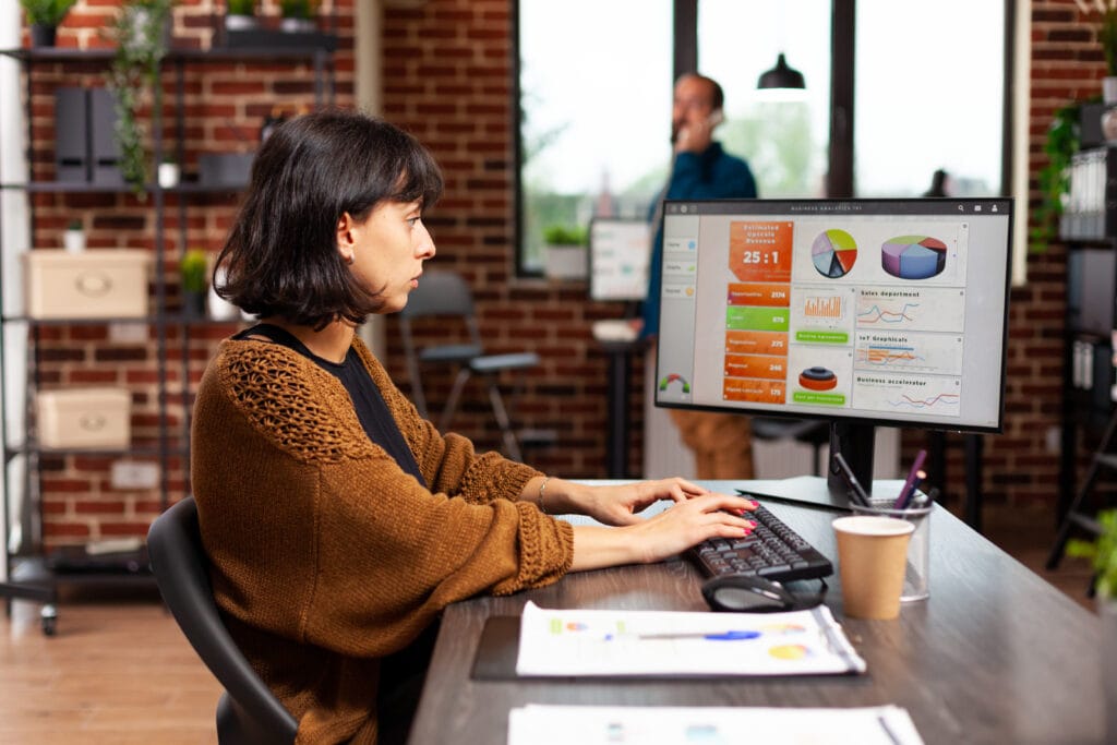 Das Foto zeigt eine Geschäftsfrau in einer Marketingagentur, die an der Datenanalyse zur Optimierung der Customer Journey arbeitet. Sie sitzt an ihrem Schreibtisch, die Hände auf der Tastatur, und scheint auf einem großen Computerbildschirm zu tippen oder durch Informationen zu navigieren. Der Bildschirm zeigt ein buntes Dashboard mit verschiedenen Diagrammen und Grafiken, wie Torten- und Balkendiagramme, die auf Kundendaten und Geschäftsanalysen hinweisen. Sie scheint sich auf den Monitor zu konzentrieren, ihr Gesichtsausdruck verrät ihr Profil und ihre Konzentration. Das Büro hat ein urbanes Loftdesign mit freiliegenden Ziegelwänden, modernen Möbeln und offenen Regalen mit Pflanzen und Büromaterial. Im Hintergrund ist ein männlicher Kollege zu sehen, der zu einer kollaborativen Atmosphäre beiträgt. Der Raum ist gut mit natürlichem Licht ausgeleuchtet, was auf ein produktives und kreatives Umfeld schließen lässt. Auf dem Schreibtisch steht ein Pappbecher mit Kaffee, ein übliches Accessoire in einer geschäftigen Arbeitsumgebung.