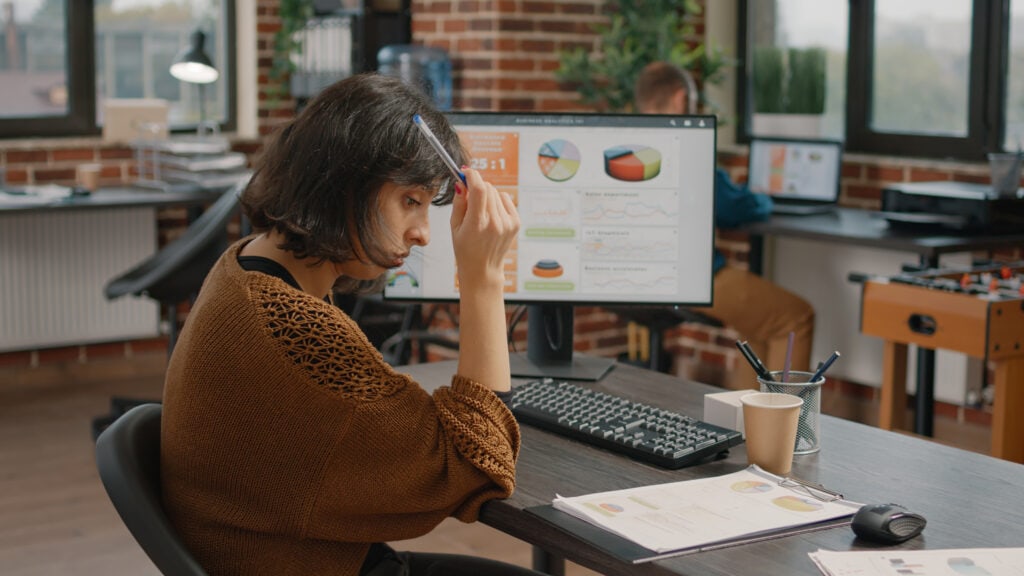 Das Foto zeigt eine nachdenkliche Geschäftsfrau in einer Marketingagentur, die tief in die Analyse von Daten zur Optimierung der Customer Journey vertieft ist. Sie sitzt an ihrem Schreibtisch und hält sich mit der rechten Hand einen Stift an die Schläfe - eine Geste, die tiefes Nachdenken oder Konzentration suggeriert. Vor ihr steht ein Monitor, auf dem eine Reihe bunter Grafiken und Diagramme zu sehen sind, die verschiedene Metriken zur Kundenbindung widerspiegeln. Zu ihrer Rechten stehen eine Tasse mit Stiften und Bleistiften, eine Papiertasse mit Kaffee und eine Maus auf einem sauberen Schreibtisch mit verstreuten Papieren, die zusätzliche Daten oder Notizen enthalten. Die Büroumgebung strahlt eine ungezwungene und doch professionelle Atmosphäre aus, mit persönlichen Akzenten wie einem Kickertisch im Hintergrund, der ein Element der kreativen und entspannten Arbeitskultur darstellt. Durch die großen Fenster fällt natürliches Licht ein, und die Inneneinrichtung wird durch freiliegende Ziegelwände ergänzt, die eine moderne und offene Arbeitsumgebung schaffen. Die Geschäftsfrau in Business-Kleidung mit Strickjacke scheint gerade eine Strategie zu entwickeln oder ein Problem zu lösen.