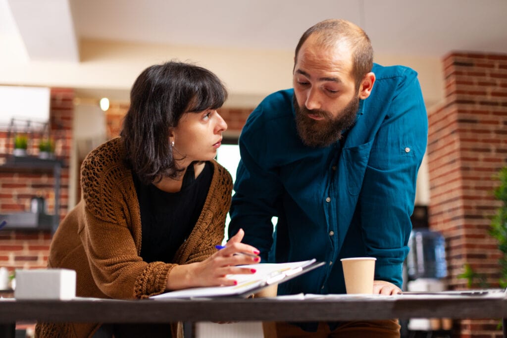 Das Foto fängt einen intensiven Moment eines Fachgesprächs zwischen einer Geschäftsfrau und ihrem männlichen Kollegen in einer Marketingagentur ein. Die Frau hält einen Bericht mit Daten zur Customer Journey in der Hand und macht eine Aussage oder stellt eine Frage. Ihr Blick ist auf ihren Kollegen gerichtet, der sich aufmerksam vorbeugt und einen Stift in der Hand hält, was darauf hindeutet, dass er sich aktiv am Gespräch beteiligt. Der Schauplatz ist ein Büroraum mit einer ungezwungenen, aber dennoch professionellen Atmosphäre, die durch die freiliegenden Ziegelwände und das moderne Mobiliar vermittelt wird. Die beiden Personen stehen vor einem Schreibtisch mit Papieren und einem Laptop, was darauf hindeutet, dass sie mitten in der Arbeit stecken. Die Frau ist leger gekleidet und trägt eine Strickjacke, während ihr Kollege ein blaues Hemd trägt, was auf einen eleganten und freundlichen Arbeitsstil hindeutet. Das Foto suggeriert ein kollaboratives und dynamisches Arbeitsumfeld, in dem der Austausch von Ideen und die Entwicklung von Strategien integraler Bestandteil des Arbeitsprozesses sind.