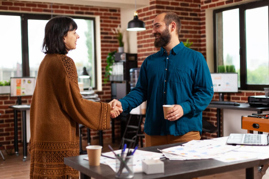 Das Foto zeigt eine Geschäftsfrau und einen Geschäftsmann, die sich die Hände schütteln, was eine herzliche und freundliche Begrüßung oder eine Vereinbarung in einer Marketingagentur zu sein scheint. Beide lächeln, was auf eine positive Interaktion hindeutet und möglicherweise den erfolgreichen Abschluss eines Gesprächs oder den Beginn einer Partnerschaft signalisiert. Die Frau trägt eine braune Strickjacke, der Mann ein blaues Button-Down-Hemd, was auf eine professionelle, aber entspannte Büroumgebung hindeutet. Im Hintergrund ist ein Büro mit freiliegenden Backsteinwänden, großen Fenstern und moderner Designästhetik zu sehen. Auf dem Schreibtisch liegen verschiedene Marketingmaterialien und Dokumente, darunter Grafiken und Diagramme, die vermutlich zur Analyse und Optimierung der Customer Journey verwendet werden. Die Umgebung ist gut ausgeleuchtet, natürliches Licht fällt durch die Fenster, und das Büro ist mit modernen Möbeln und Büropflanzen ausgestattet, die eine einladende und produktive Atmosphäre schaffen. Der Händedruck in der Mitte des Fotos zeigt einen Moment der professionellen Kameradschaft und der Zusammenarbeit in der Agentur.