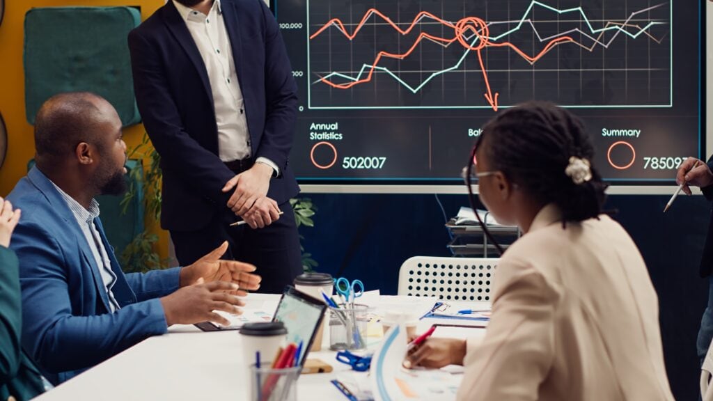 Ein Team diskutiert intensiv über Marketing-Daten in einem Meetingraum. Im Hintergrund zeigt ein großer Bildschirm ein Jahresreport-Diagramm mit Finanz- und Marktstatistiken. Ein Geschäftsmann im blauen Anzug erklärt eine Strategie, während Kollegen aufmerksam zuhören und Notizen machen.