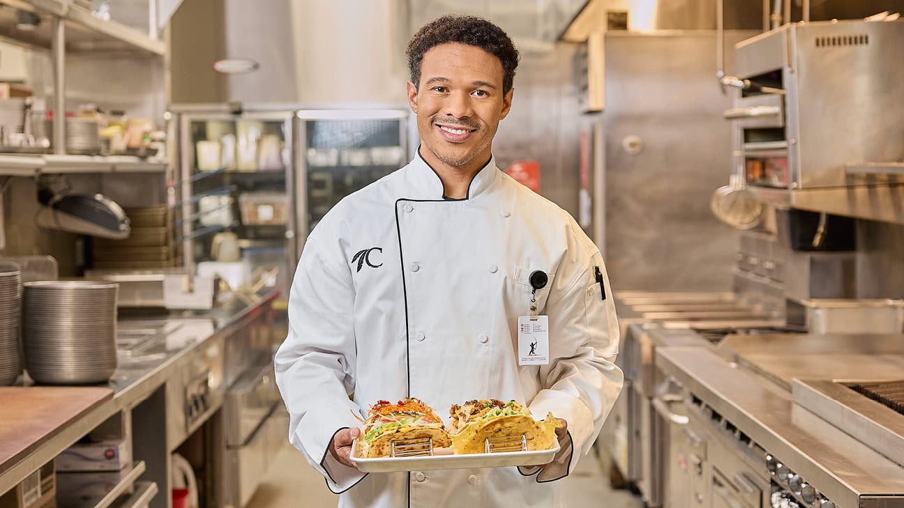 a man with curly brown hair and a chef's uniform smiles in a kitchen.