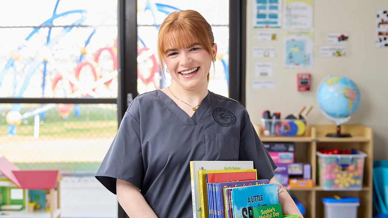 A red-headed, female woman in scrubs smiles in a classroom.