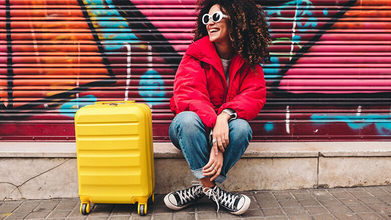 Bright young woman with a yellow suitcase sitting against colorful graffiti wall, travel or lifestyle concept.
