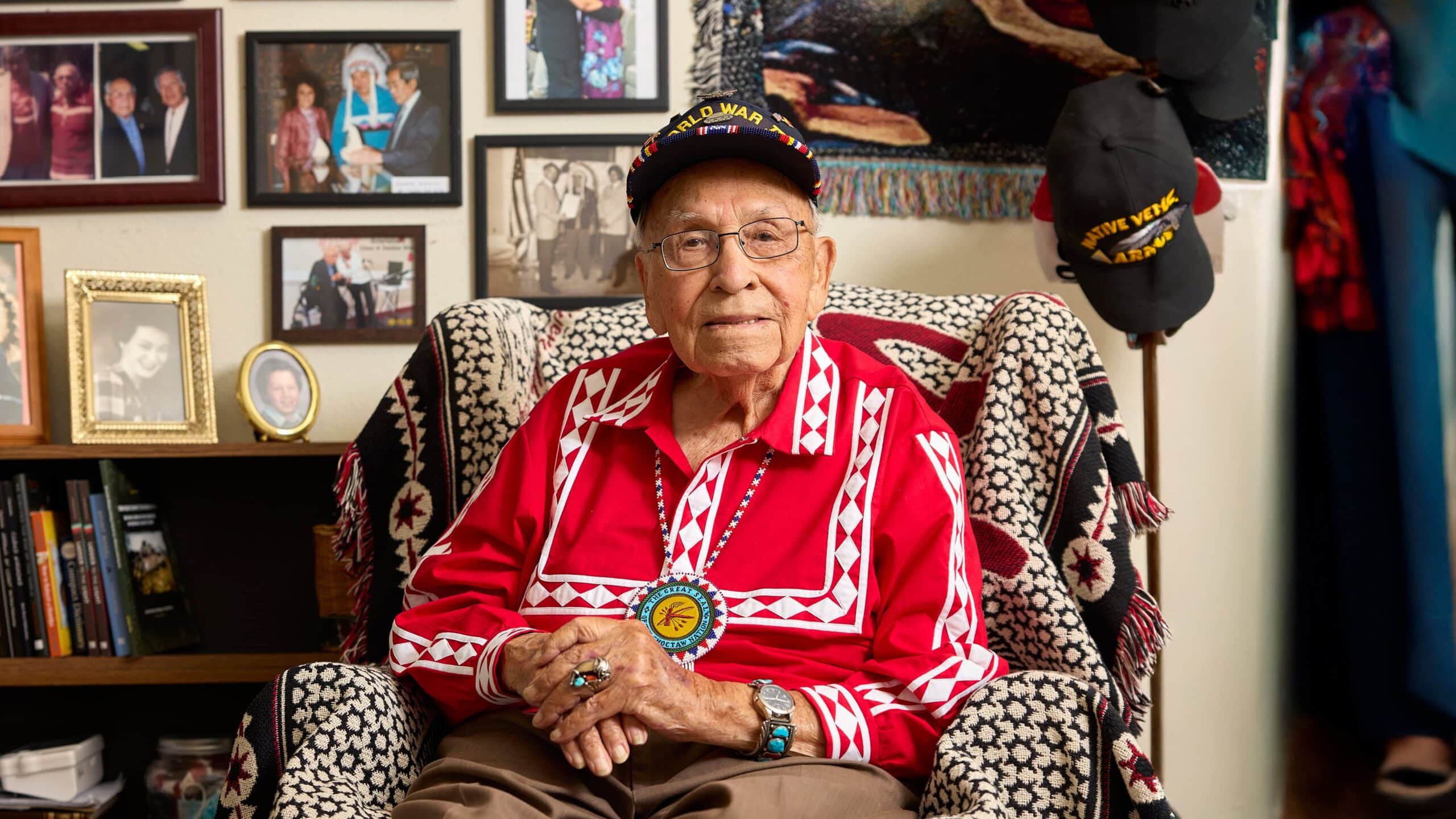 Profile image of Choc sitting in a chair in his Choctaw attire with a medallion around his neck. His army cap in the background.