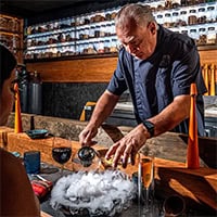 Man behind bar pours liquid into a bowl that produces smoke.