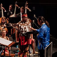 A man in Indigenous clothing stands in front of an orchestra, and a conductor in a blue traditional shirt.