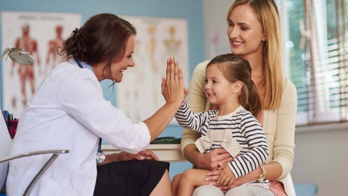 A female doctor giving a high-five to a smiling young girl sitting on her mother’s lap during a medical appointment. The mother looks on happily in a bright, welcoming exam room with anatomy posters on the wall.