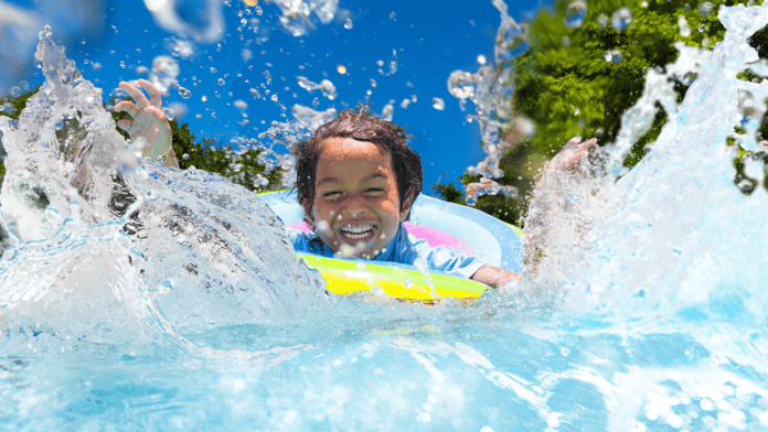 A joyful young child splashing in a pool while floating on a colorful inflatable ring. The child is laughing with arms raised as water sprays up around them under a bright blue sky.