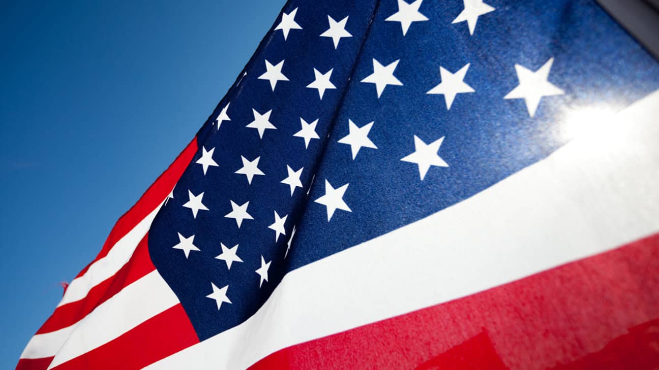 Close-up of the American flag waving in the wind against a clear blue sky, showing white stars on a blue field and red and white stripes.