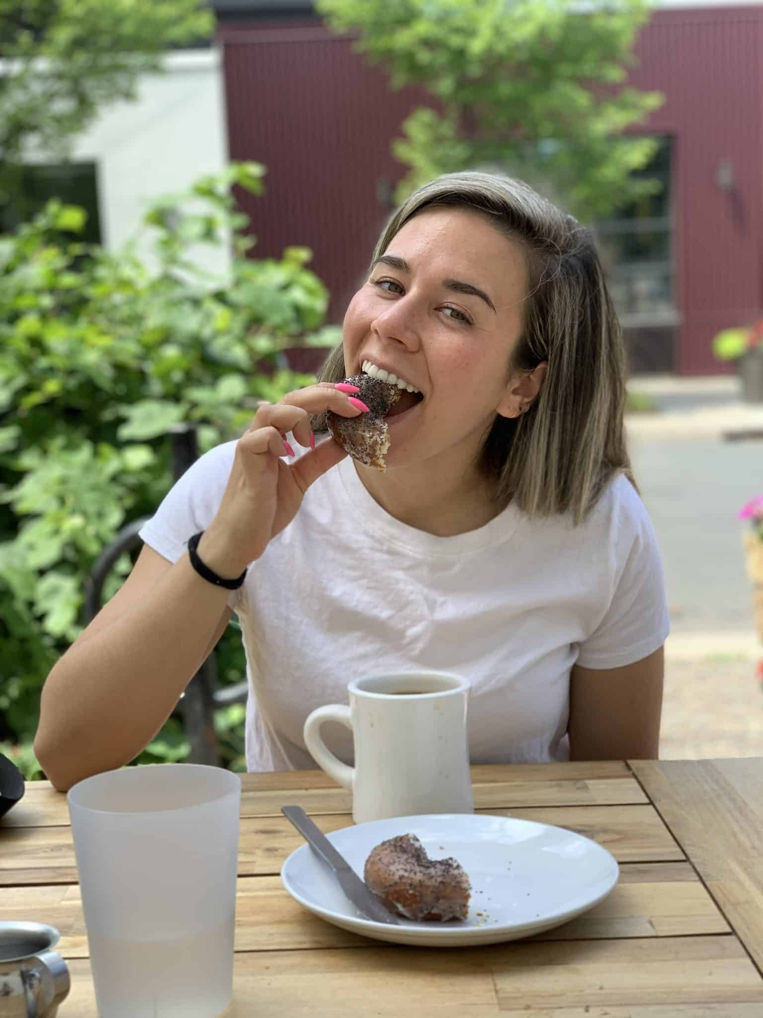 I'm sitting a table outside, happily eating a donut. I have a white t-shirt on and a mug of coffee on the table in front of me.
