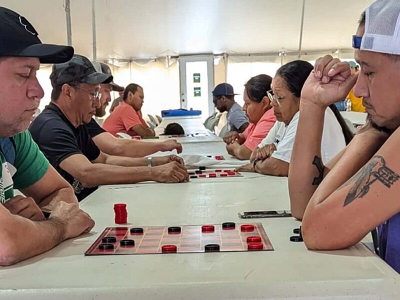 A man in a green shirt and a man in a purple shirt look down at their checkerboard during a game of checkers.
