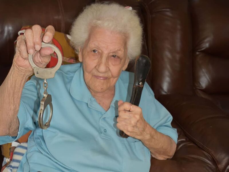 An old woman in a blue blouse holds up handcuffs and a flashlight.