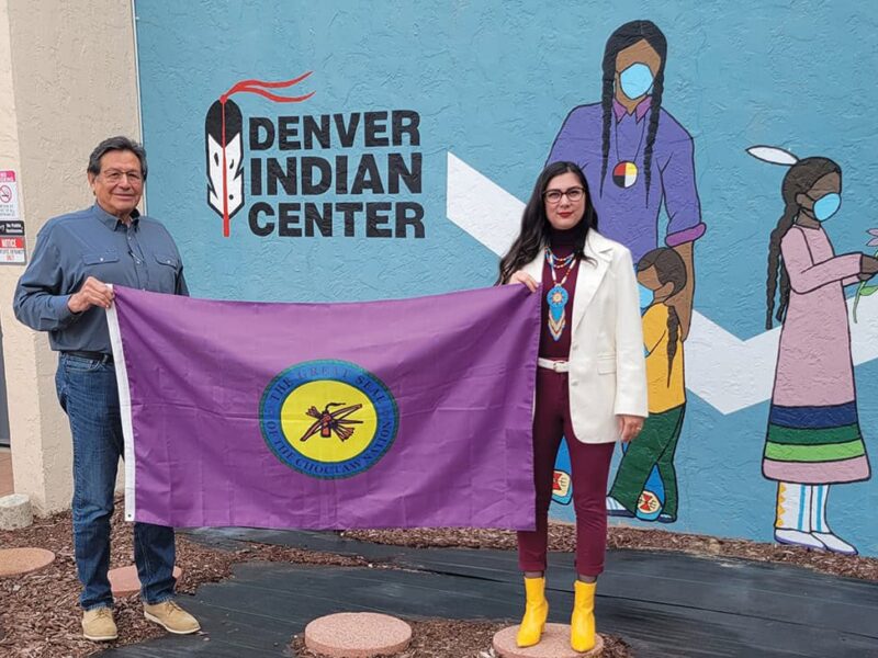 Two people stand in front of the Denver Indian Center holding the Choctaw Nation of Oklahoma flag
