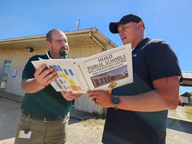 Two man look at a book containing spill guidelines for Hugo Public Schools.