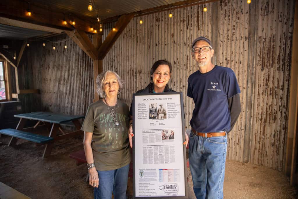 Three people pose for a photo with a display about the Choctaw code talkers.