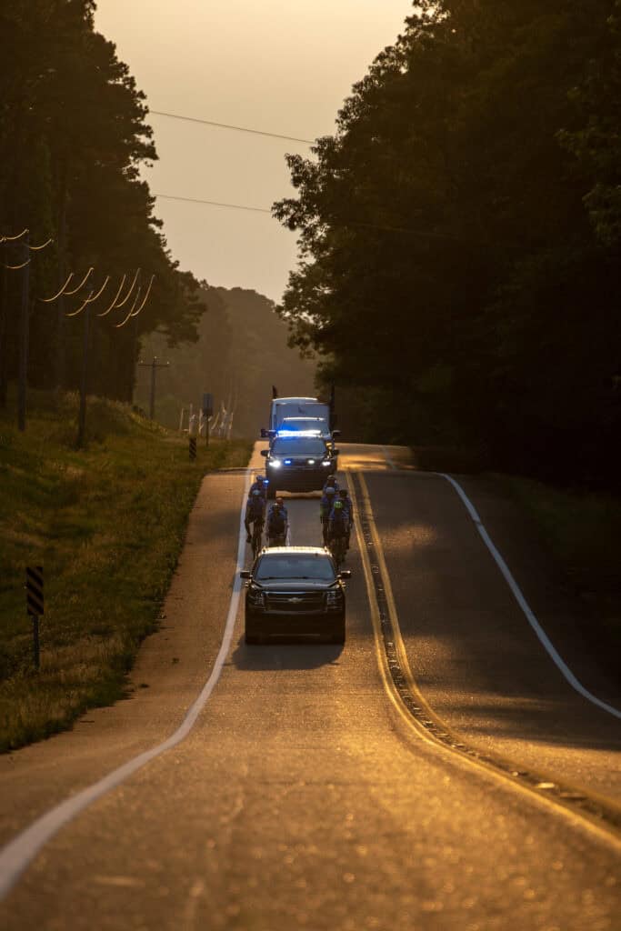 Cyclists ride down the road escorted by safety vehicles as the sun rises.