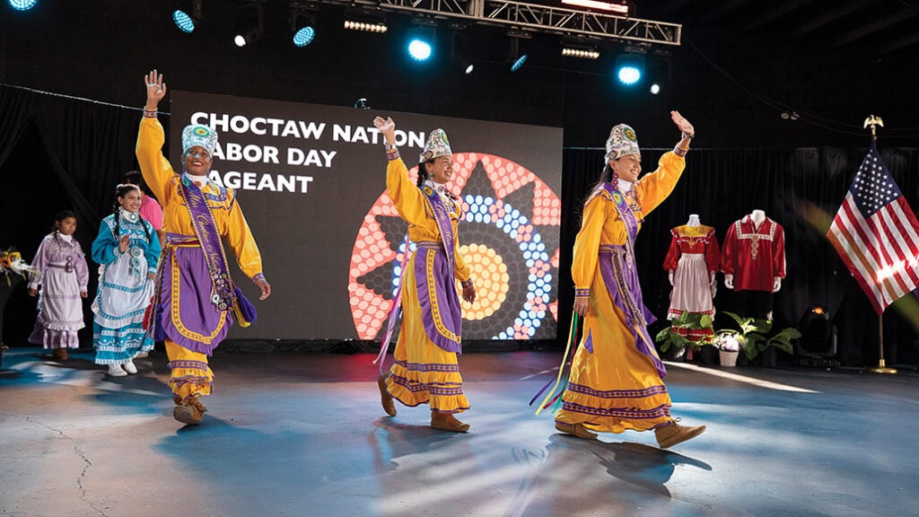 Three young women walk on stage wearing traditional regalia.