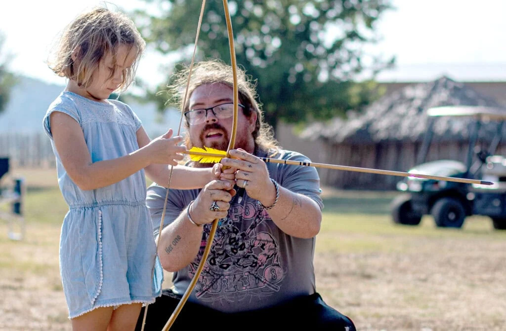 A man helps a young woman place an arrow on a bow.