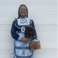 Young girl stands, holding feathers and a basket, wearing a blue and white traditional dress.