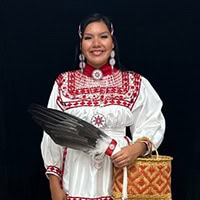 Young woman stands, holding a feather and basket, wearing a white and red traditional dress.