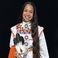 Young girl stands, holding feathers, wearing a white and orange traditional dress.