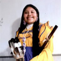 Young girl stands, holding feathers and a flute, wearing a yellow and blue traditional dress.