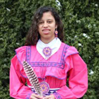 Young girl stands, holding a feather, wearing a bright pink and purple traditional dress.