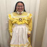 Young girl stands, smiling, wearing a yellow and white traditional dress.
