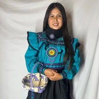 Young girl stands, holding a basket, wearing a turquoise and black traditional dress.
