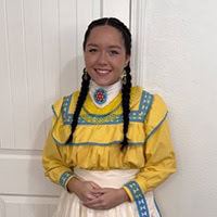 Young woman stands, smiling, wearing a yellow, blue and white traditional dress.