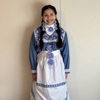 Young girl stands, smiling, wearing a blue and white traditional Choctaw Dress.