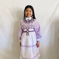Young girl stands, smiling, wearing a lilac and white traditional Choctaw dress.