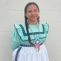 A young girl stands, wearing a teal, navy blue and white traditional dress.