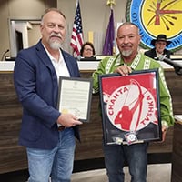 Man in blue blazer holds up plaque, while councilmember Anthony Dillard holds up a framed choctaw veteran's flag.