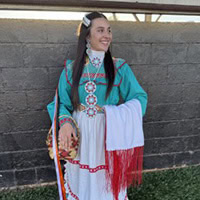 Young girl stands, holding a shaw and basket, wearing a teal, white and red traditional dress.