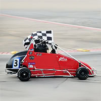 A young boy holds a checkered flag as he drives his race cart.