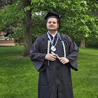A man poses fr the camera wearing a graduation gown.