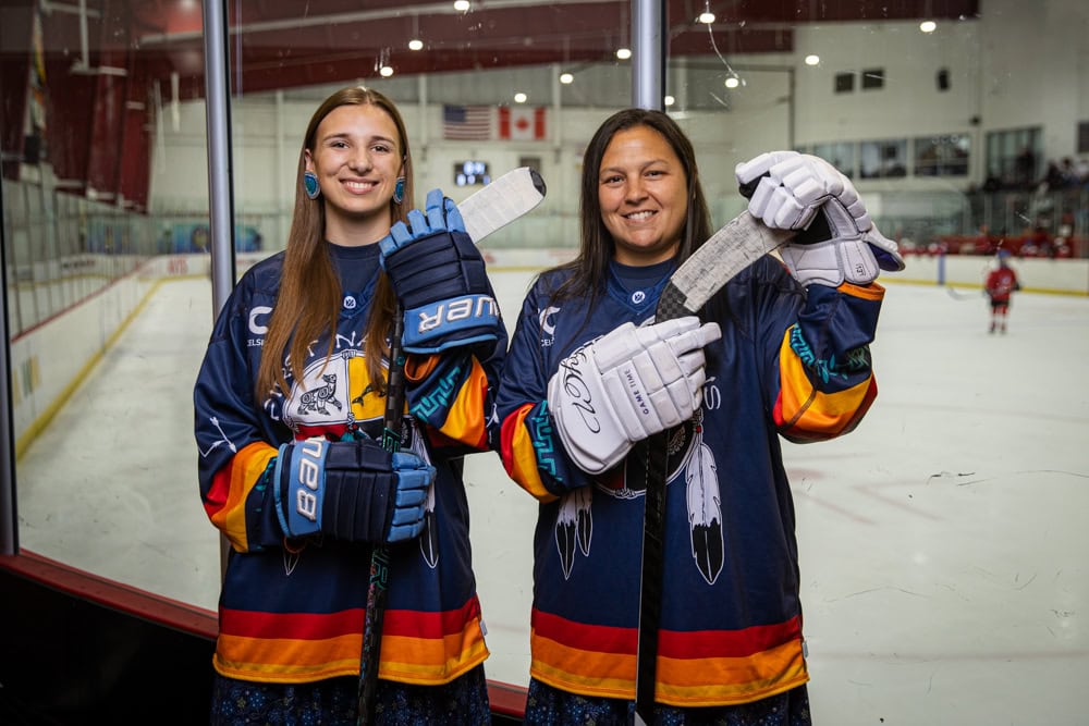 Two hockey players pose for a photo, holding their sticks, in front of the rink.
