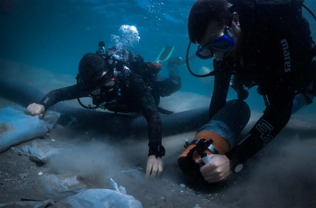 Two people in scuba suits use large hose on ocean floor to sift through sand.