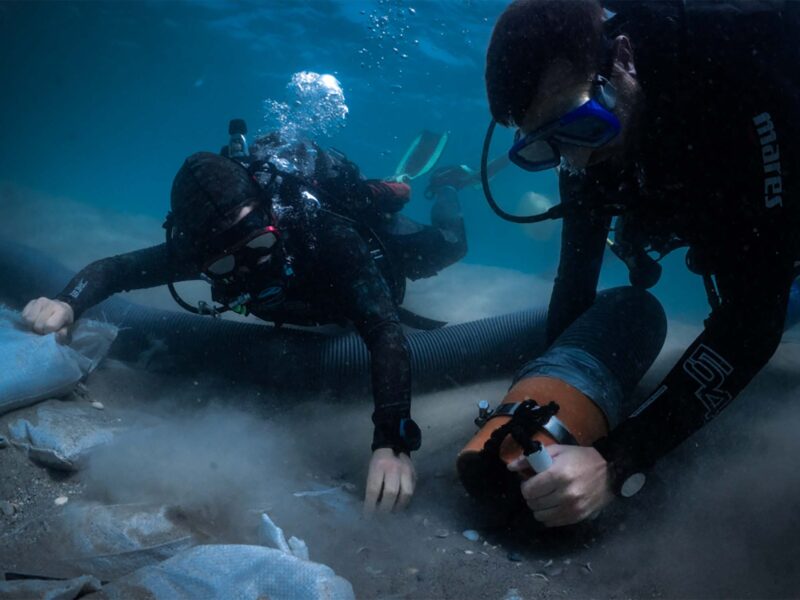 Two people in scuba suits use large hose on ocean floor to sift through sand.