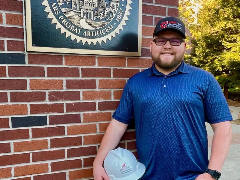 Man stands in front of brick wall holding a hard hat.