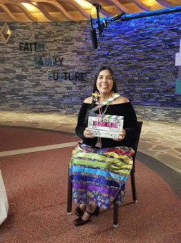 A woman sits in a chari wearing modern Native inspired clothing.