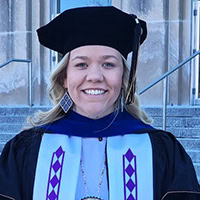 Young woman dressed in doctoral cap and gown smiles.