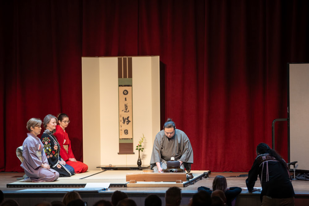 four people on a stage in traditional Japanese dress perform a tea ceremony as a Native man illustrates it.