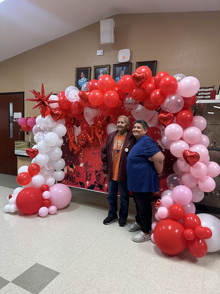 Two people pose for a photo surrounded by red, pink and white balloons. 