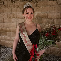 A young woman poses for a photo wearing a pageant crown and ribbon.